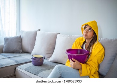 Woman In Raincoat Waiting For Plumber To Fix Flood. Frustrated Female Client In Raincoat Holding Pot While Water Leaking From Roof In Hotel. Woman Collecting Leaking Water From Ceiling In Living Room