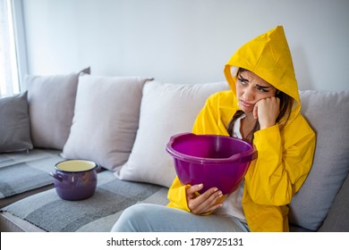 Woman In Raincoat Waiting For Plumber To Fix Flood. Frustrated Female Client In Raincoat Holding Pot While Water Leaking From Roof In Hotel. Woman Collecting Leaking Water From Ceiling In Living Room