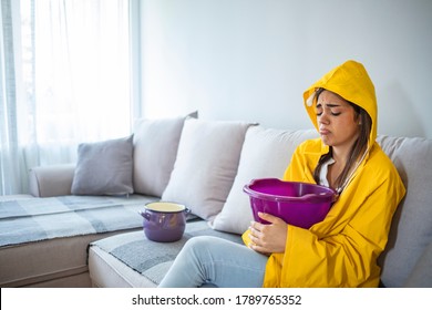 Woman In Raincoat Waiting For Plumber To Fix Flood. Frustrated Female Client In Raincoat Holding Pot While Water Leaking From Roof In Hotel. Woman Collecting Leaking Water From Ceiling In Living Room