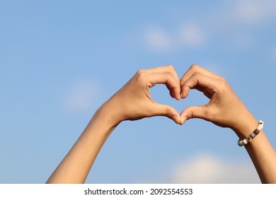 A woman raises her hand above her head to make a heart symbol on the background of the bright morning sky because the heart symbol signifies love, friendship and kindness to one another.