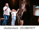 A woman raises her hands in worship during a church service, facing the worship leaders on stage. Capturing a moment of faith, praise, and spiritual connection in a vibrant worship environment.