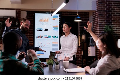 Woman Raising Hand To Ask A Question In Late Night Business Meeting With Sales Department In Startup Office. Coworker Interrupting Manager Presenting Analytics Charts With Asian Coworker.