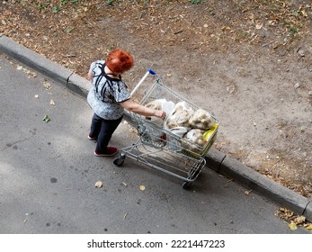 A Woman Reaches Out To A Package Of Potatoes In The Cart. View From Above. Fallen Leaves Are Visible On The Pavement.