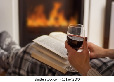 Woman Reading Book And Drinking Red Wine In Front Of The Fireplace. Girl Warming On Fire Flame And Covered Legs With Blanket. Winter And Long Cold Night Concept. Close Up, Selective Focus