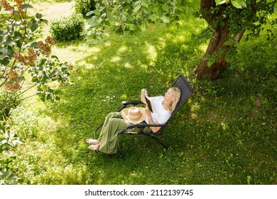 Woman Reading A Book In Garden Recliner Chair. Summer Vacation In The Yard. Mature Scandinavian Female Life In Retirement. Good Living. View From The Top. Beautiful Middle Aged Woman Resting