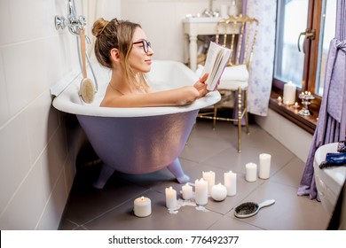 Woman Reading A Book Lying In The Retro Violet Bath In The Vintage Bathroom Decorated With Candles