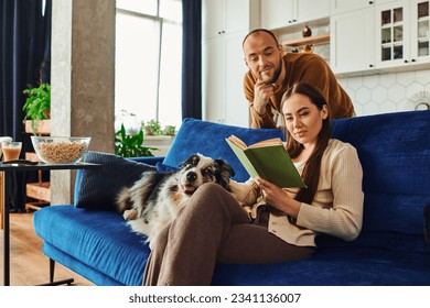 Woman reading book near boyfriend and border collie dog on vouvh in living room at home