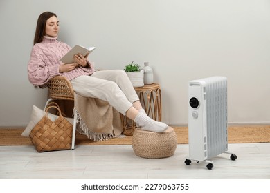 Woman reading book near modern portable electric heater indoors