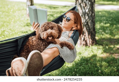 Woman reading book relaxing in hammock with her fluffy brown Maltipoo dog on sunny day. Both looking content and happy. This outdoor scene captures joy of bonding with pets and enjoying togetherness - Powered by Shutterstock