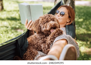 Woman reading book relaxing in hammock with her fluffy brown Maltipoo dog on sunny day. Both looking content and happy. This outdoor scene captures joy of bonding with pets and enjoying togetherness