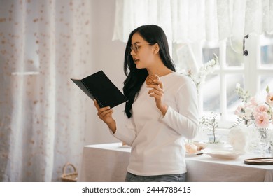 A woman is reading a book while eating a cookie. The scene is set in a kitchen with a table and a vase of flowers. The woman is wearing glasses and she is enjoying her book and snack - Powered by Shutterstock