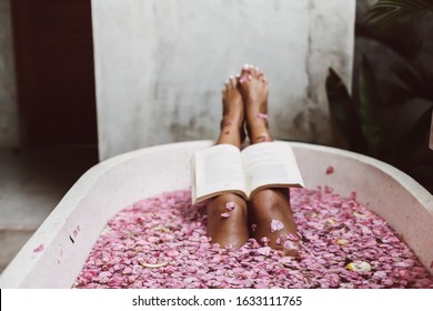 Woman Reading Book While Relaxing In Bath Tub With Flower Petals, POV. Organic Spa Relaxation In Luxury Bali Outdoor Bath.