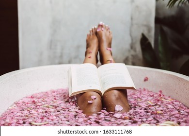 Woman reading book while relaxing in bath tub with flower petals, POV. Organic spa relaxation in luxury Bali outdoor bath.