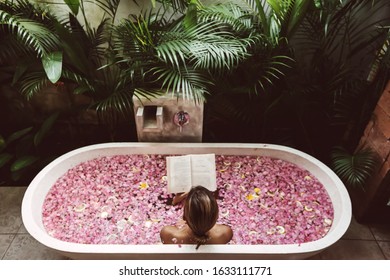 Woman reading book while relaxing in bath tub with flower petals. Organic spa relaxation in luxury Bali outdoor bath.