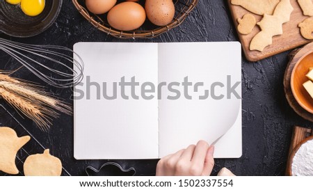 Woman is reading cookbook recipe of making Halloween cookies with baking ingredients, design concept of cooking class, top view, flat lay, overhead.