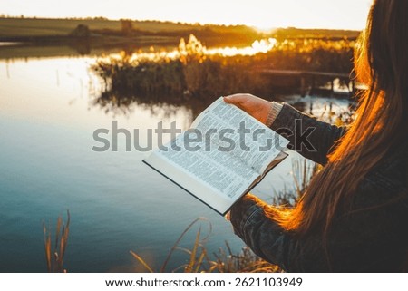 Woman reading open Bible by a peaceful lake during golden sunset, with warm light reflecting on water and long hair glowing in sunlight. Tranquil outdoor moment of faith and reflection.