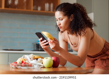 Woman Reading Phone, Researching A Diet With Fresh Fruit While Relaxing In A Kitchen At Home. Young Female Searching For A Recipe, Cleanse Or Detox Online. Lady Checking Nutritional Value Of A