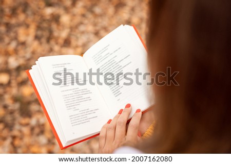 Woman reading poems in autumn park. Back view of opened book with red cover in hands of a girl.