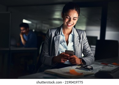 Woman Reading Text Late On Phone, Smiling At A Message And Looking At Social Media Posts Online While Working In A Dark Office At Night. Happy Corporate Worker Checking Emails And Doing Overtime