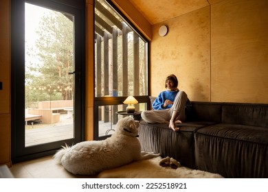 Woman reads a book while sitting relaxed on a couch by the window with her dog at wooden cottage in forest. Concept of home comfort and rest in a house on nature - Powered by Shutterstock