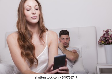 woman reads message on smart phone in bed while man looks jealous