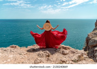 A woman, rear view in a red flying dress fluttering in the wind, a girl in a fluttering dress on the background of the sea. A straw hat hangs at the back of the neck.