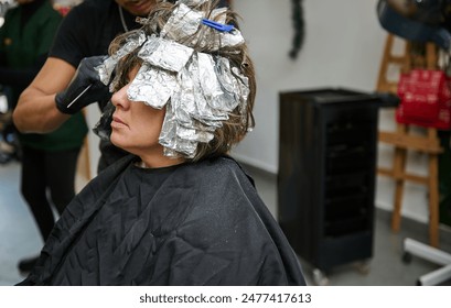 A woman receiving a hair coloring treatment with aluminium foils at a beauty salon. Professional hairstylist applying dye. - Powered by Shutterstock