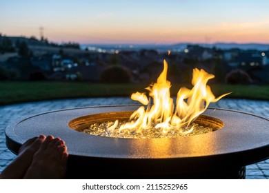 A woman reclines on a hillside patio terrace with her feet up on the edge of a flaming fire pit as the sun sets and the city lights go on in the distant view.