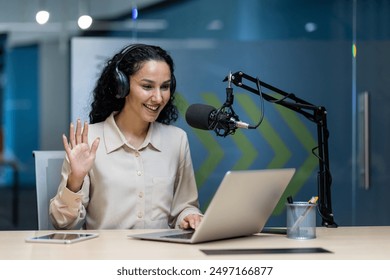 Woman recording podcast in modern office setup wearing headphones, speaking into microphone. Professional podcasting environment with laptop and tablet on desk. Happy and engaged expression - Powered by Shutterstock