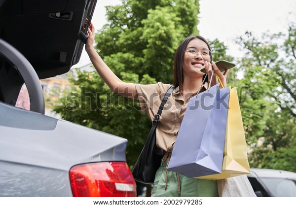 Woman recording voice message and standing\
near her car while holding shopping\
bags