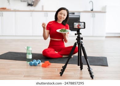 Woman in red activewear recording healthy eating video with smartphone in modern home kitchen. Demonstrating healthy lifestyle, balanced diet, and fitness goals during video. - Powered by Shutterstock