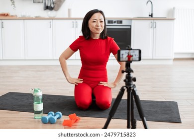 Woman in red activewear recording home workout tutorial on smartphone. Setup includes exercise mat, dumbbells, water bottle, and resistance bands in bright, modern kitchen. - Powered by Shutterstock