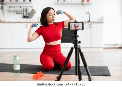 Woman in red athletic outfit exercising with dumbbells in home kitchen. Filming workout session with smartphone on tripod for online audience. Healthy lifestyle, fitness, and home workout concept. - Powered by Shutterstock