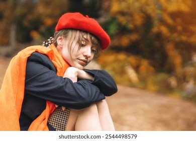 A woman in a red beret and orange shawl sits thoughtfully among autumn leaves in a serene outdoor setting - Powered by Shutterstock