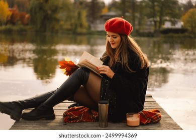 Woman in a red beret reading book on wooden pontoon. Autumn season. Female with trendy clothes relaxing near water reading book and drink coffee. Autumn mood. Autumn concept. Autumn day. - Powered by Shutterstock