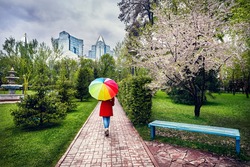 Woman In Red Coat With Rainbow Umbrella Walking In The Park With Blooming Trees At Overcast Sky