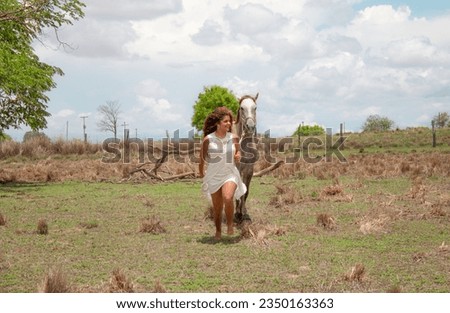 Woman with red curls and white dress, with horse in a green field and sky with clouds.