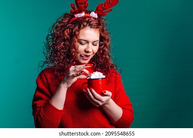 A Woman With Red Curly Hair In A Red Jacket And Deer Horns On Her Head. The Girl Is Holding A Red Mug With Marshmallow In Her Hands And Stuck Out Her Tongue In Anticipation Of Yummy. Christmas 