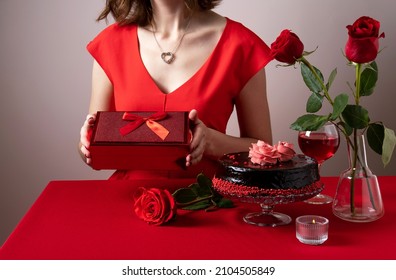 Woman In Red Dress Hand Holding A Red Gift Box. Valentines Day Dinner With Table Place Setting With Red Wine, Candles And Chocolate Cake. Romantic Celebration Of Valentine's Day. 