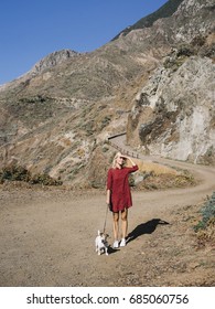 Woman in red dress with Jack Russell Terrier posing in sunlight on background of road in mountains. 