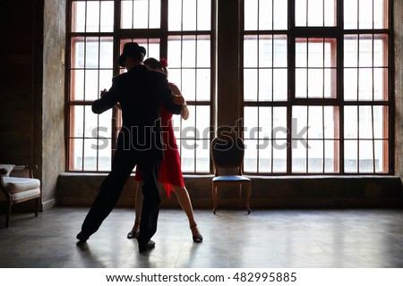 Woman in red dress and man in black suit dance tango near big window in room
