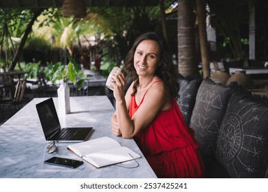 Woman in a red dress smiling and holding a coffee cup at an outdoor workspace with a laptop, notebook, and tropical plants. Relaxed, inspiring setting with a cozy, natural atmosphere - Powered by Shutterstock