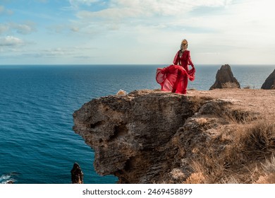 A woman in a red dress stands on a rocky cliff overlooking the ocean. The scene is serene and peaceful, with the woman's flowing dress adding a sense of grace and beauty to the landscape. - Powered by Shutterstock