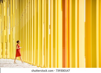 Woman In Red Dress Walking On The Modern Yellow Building Wall Background. General Plan With Abstract Geometric Composition