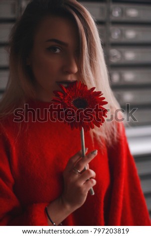 woman with a red flower - gerbera on the background of postal cells. she is dressed in a red top or sweater. hair dyed ombre, blonde and brunette. girl with makeup.