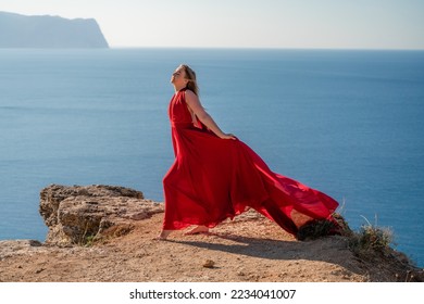 A woman in a red flying dress fluttering in the wind, against the backdrop of the sea. - Shutterstock ID 2234041007