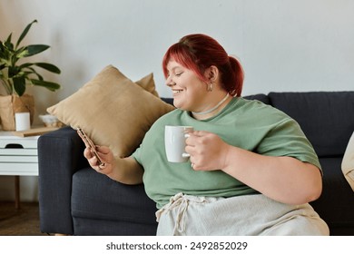 A woman with red hair sits on a couch, smiling as she looks at her phone and sips from a mug. - Powered by Shutterstock