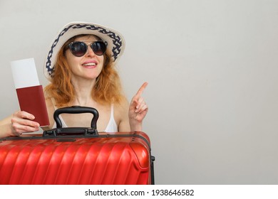 Woman with red hair smiling, holding a passport with tickets, standing with a suitcase and pointing her finger at a light background