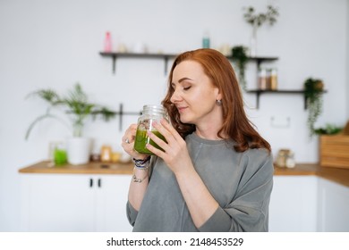 A Woman With Red Hair Stands In Her Kitchen And Sniffs Celery Juice In A Glass