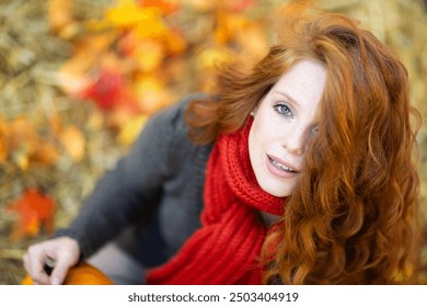 A woman with red hair, wearing a gray sweater and red scarf, gazes upward with a thoughtful expression, surrounded by vibrant autumn leaves and straw in an outdoor setting
 - Powered by Shutterstock